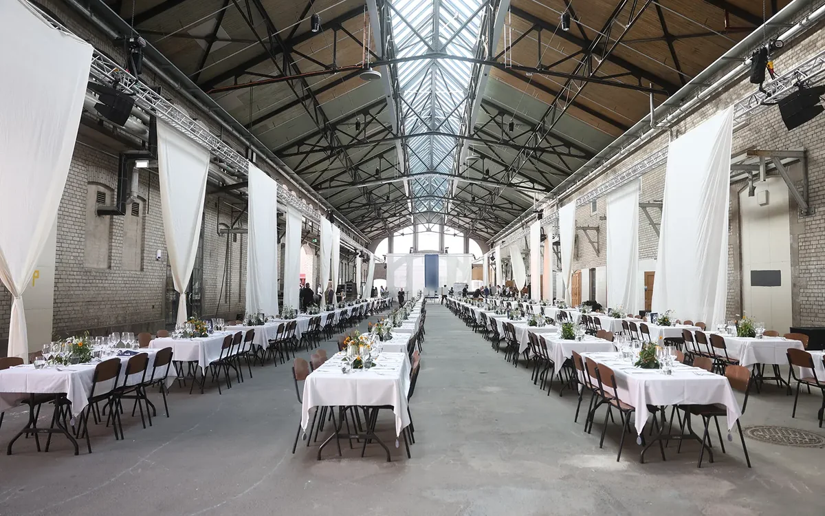 Long tables laid for a dinner in a skylit industrial hall, white drapes along the walls