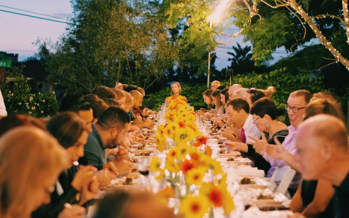 A long table of guests under string lights on a summer evening, sunflowers down the middle